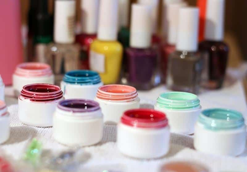 Eight small pots of nail polishes of various colors on a white table with lots of nail polish bottles in the background.