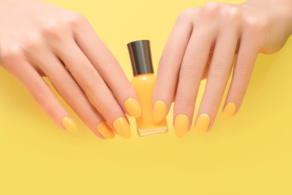 close up of a woman's hands holding onto a yellow bottle of nail polish behind a yellow background. Her fingernails are painted yellow.