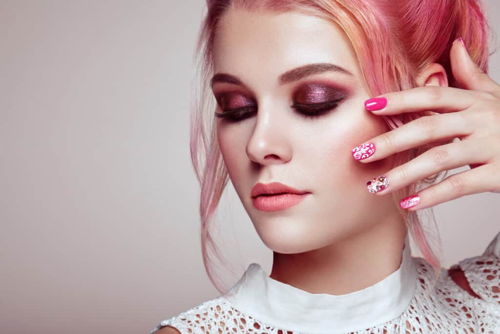 A woman is seen with closed eyes. She has pink hair, pink eyeshadow on and her hand is covering one side of her face.