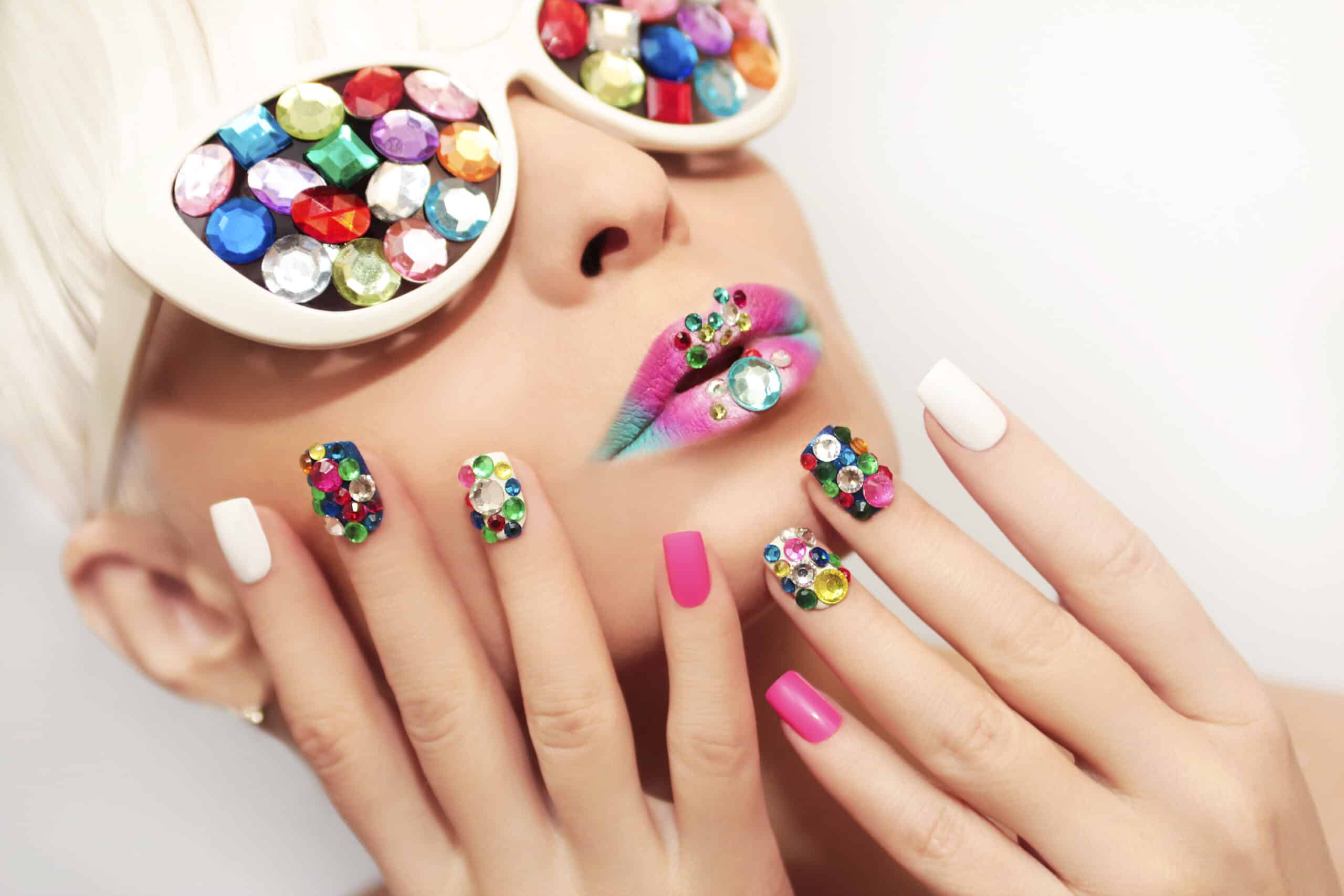A woman with white sunglasses covered in gem stones look up towards the sky. Her lips are multi-colored and covered in gems and her nails are white and pink, also covered in gems.