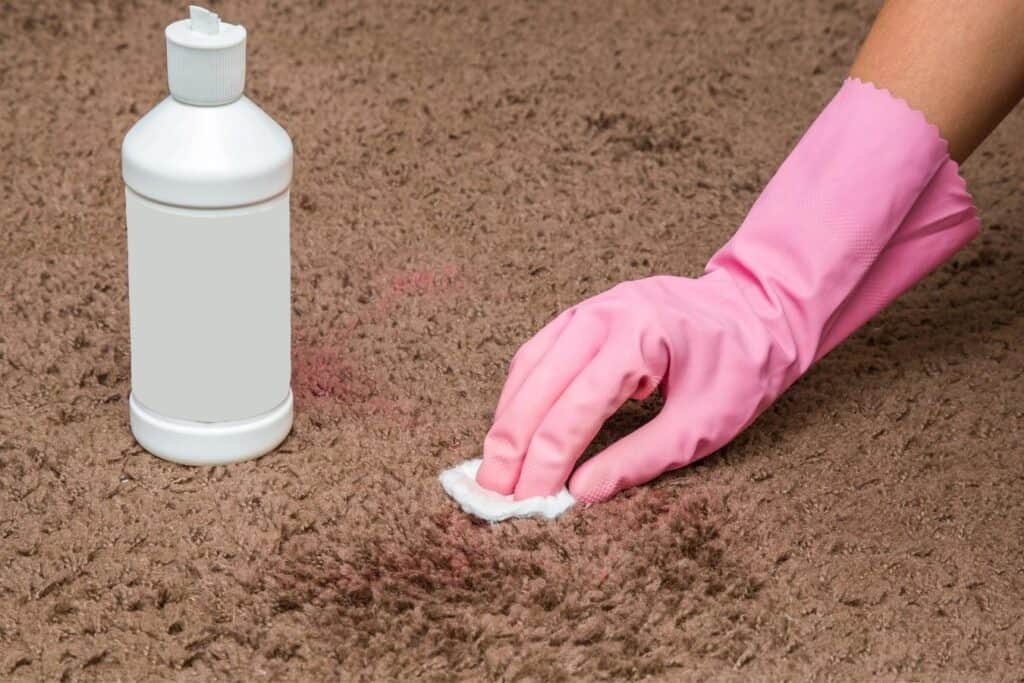 A person wearing pink gloves is removing stains from carpet. A white bottle is seen next to the hand.